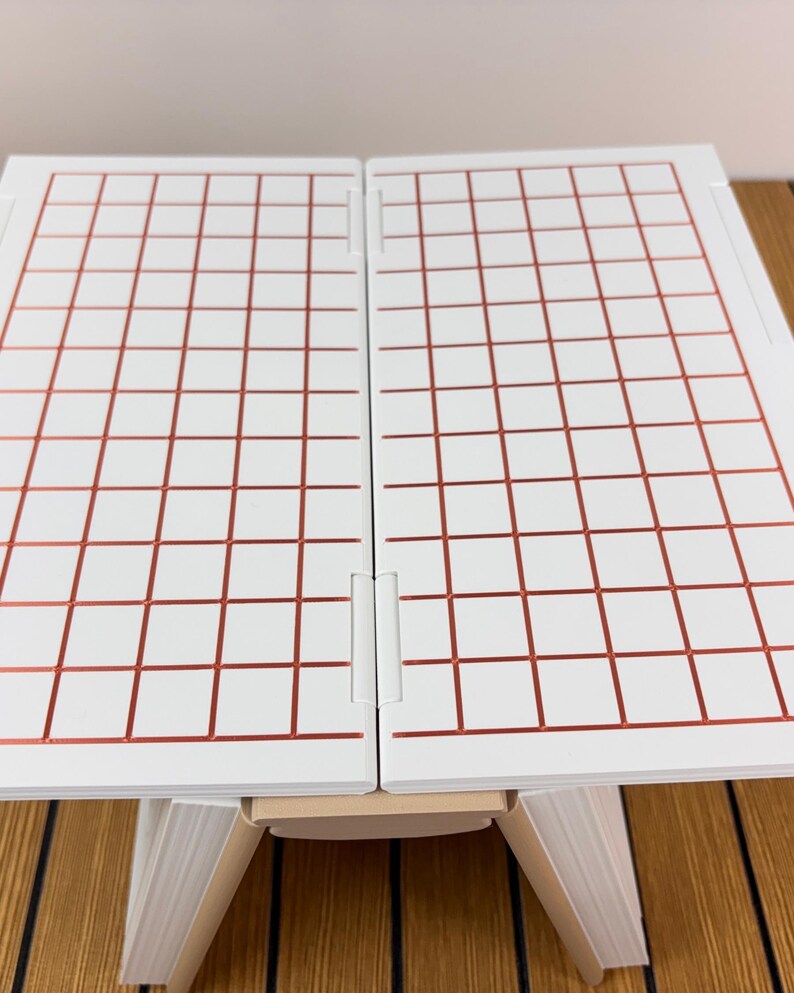 May include: A white folding board featuring a grid pattern of thin, metallic copper-colored lines. The board is composed of two rectangular panels connected by a central hinge, supported by a light-colored base structure underneath.