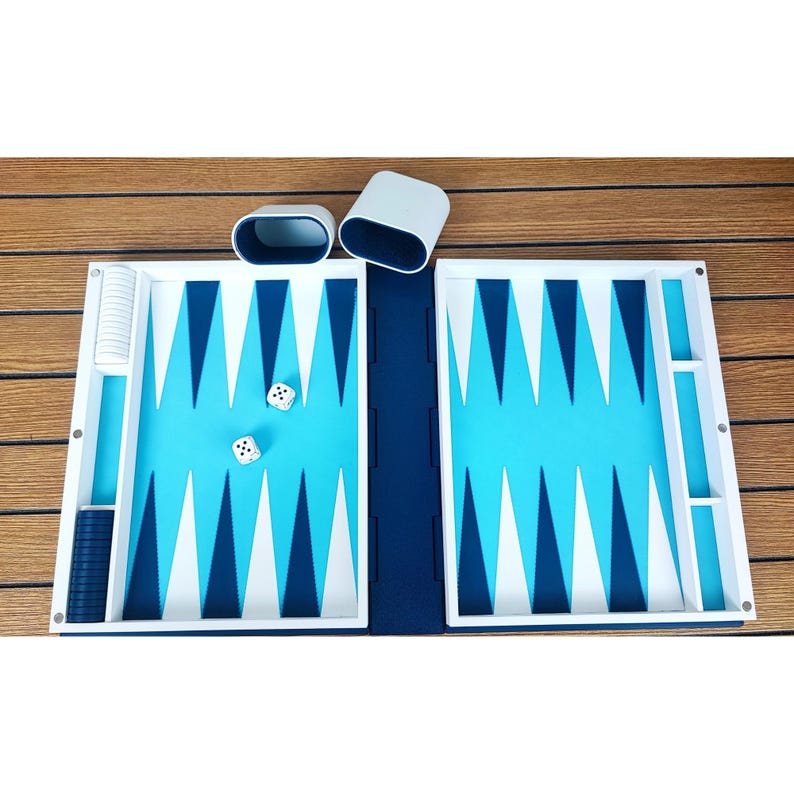 May include: A backgammon set with a blue and white playing surface. The board is open, revealing the triangular points and dice. Two white dice with black dots are on the board, and two white dice cups are nearby. The set is on a wooden surface.