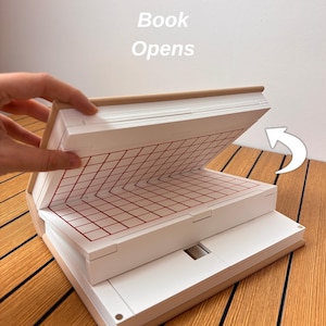 May include: A beige book-shaped object with a white interior featuring a red grid pattern on the top page. The text 'Book Opens' is printed above the object, and a white curved arrow points to the right to indicate the opening motion.
