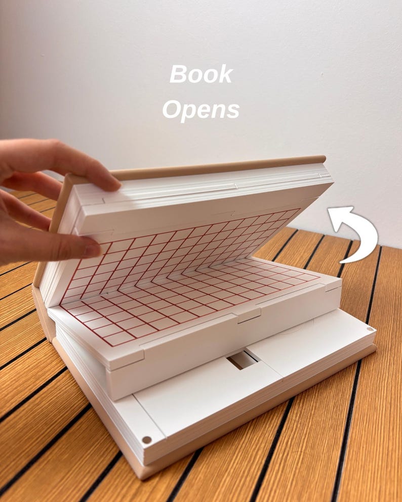 May include: A beige book-shaped object with a white interior featuring a red grid pattern on the top page. The text 'Book Opens' is printed above the object, and a white curved arrow points to the right to indicate the opening motion.
