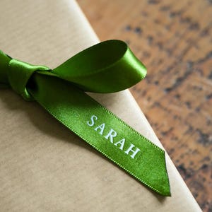 May include: A rectangular package wrapped in plain brown paper, tied with a vibrant green satin ribbon. The ribbon is tied in a bow, and the name SARAH is printed in white capital letters along one of the ribbon tails extending from the knot.