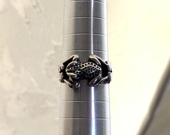 May include: A silver-toned ring featuring a frog design. The frog is detailed with textured skin and a perforated belly. The ring is displayed on a silver ring sizer, showcasing its size and shape. The background is a soft, neutral color.