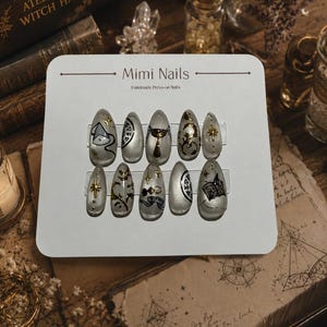 May include: A set of ten almond-shaped press-on nails on a white display card labeled Mimi Nails Handmade Press-on Nails. The clear nails feature various black and gold metallic designs including stars, celestial symbols, scrolls, and a book illustration.