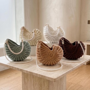 May include: Five handmade crochet handbags in various colors: sage green, beige, white, tan, and dark brown. Each bag features a unique shell-like design, a pearl strap, and a handle. The bags are displayed on a marble-topped table.