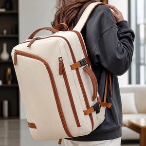 May include: Cream-colored backpack with brown leather accents. The bag features a rectangular shape with a top handle, a front zipper pocket, and adjustable straps. Brown leather straps and buckles secure the bag's front. The backpack is designed for travel or everyday use.