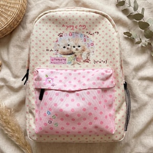 May include: A cream-colored backpack with pink polka dots featuring a graphic of two kittens in cups. The front pocket is pink with darker pink polka dots and small paw print stickers. The bag includes black zippers and a black mesh side pocket.