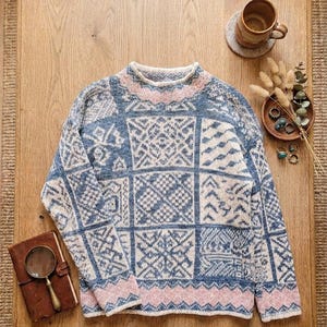Può includere: Un maglione a maniche lunghe caratterizzato da un design patchwork con motivi geometrici blu, bianchi e rosa tenue. Il capo in maglia presenta uno scollo rotondo e un bordo ripetuto a rombi e chevron lungo l'orlo e i polsini.