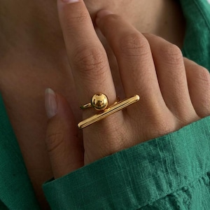 May include: A gold-toned ring featuring a smooth, polished finish. The design consists of a thin, horizontal bar element positioned parallel to a small, spherical ball bead, creating a geometric and open-ended structure on the finger.