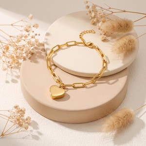 Könnte beinhalten: Ein goldfarbenes Armband mit einer Kette aus länglichen, rechteckigen Gliedern und einem einzelnen, glatten, massiven herzförmigen Anhänger. Das Armband verfügt über eine feine Verlängerungskette mit einem kleinen Karabinerverschluss.