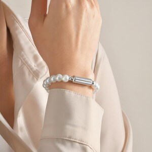 Op de afbeelding: Een armband bestaande uit ronde witte parels en een gladde, cilindervormige sluiting van zilverkleurig metaal. De parels zijn uniform van grootte en gerangschikt in een enkele streng, verbonden door het metalen onderdeel als sluiting.