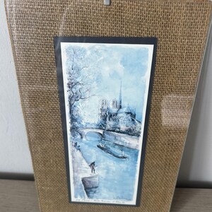 May include: A rectangular framed print featuring a blue-toned watercolor painting of the Notre-Dame Cathedral in Paris, situated along the Seine River with a bridge and a boat, set against a textured brown burlap matting within a clear glass cover.