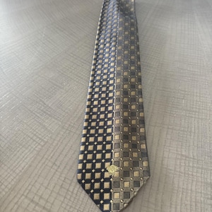 May include: A patterned necktie with a geometric design in navy blue, gold, and gray. The tie features a repeating square pattern and a gold emblem near the tip. The tie is made of a smooth, possibly silk, material.
