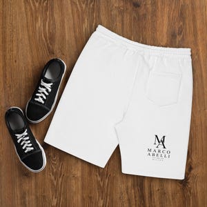 Peut inclure: Short blanc avec taille élastique et poche arrière, accompagné de baskets en toile noires. Le short porte le logo "M MARCO ABELLI MILANO" en noir. Les baskets ont des lacets et des semelles blanches.