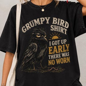 以下が含まれることがあります： 不機嫌そうな鳥のグラフィックと「GRUMPY BIRD SHIRT, I GOT UP EARLY, THERE WAS NO WORM」というテキストがプリントされた黒の半袖Tシャツ。デザインには小さな太陽のアイコンと地面にいる虫が黄色と白のインクで描かれています。
