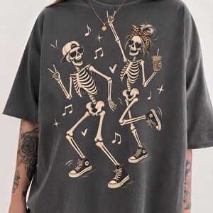 May include: A charcoal grey short-sleeved t-shirt featuring a graphic print of two dancing skeletons. One skeleton wears a baseball cap and sunglasses, while the other wears a leopard print headband and heart-shaped sunglasses, holding a drink.