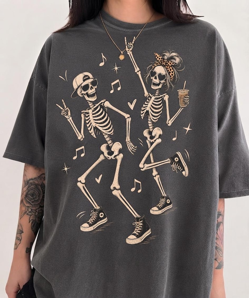 May include: A charcoal grey short-sleeved t-shirt featuring a graphic print of two dancing skeletons. One skeleton wears a baseball cap and sunglasses, while the other wears a leopard print headband and heart-shaped sunglasses, holding a drink.