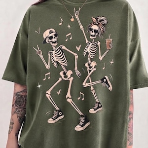 May include: An olive green t-shirt featuring a graphic print of two dancing skeletons. One skeleton wears a baseball cap and sunglasses, while the other wears a leopard print headband and heart-shaped sunglasses, holding a drink with a straw.