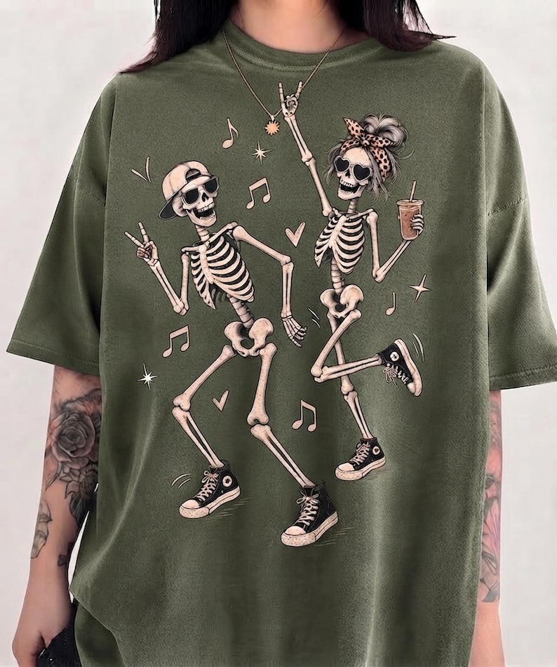 May include: An olive green t-shirt featuring a graphic print of two dancing skeletons. One skeleton wears a baseball cap and sunglasses, while the other wears a leopard print headband and heart-shaped sunglasses, holding a drink with a straw.