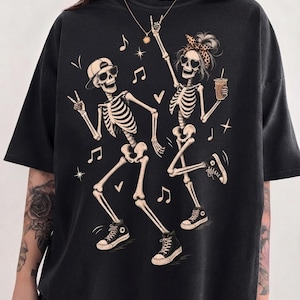 May include: A black short-sleeved t-shirt featuring a graphic print of two dancing skeletons. One skeleton wears a baseball cap and sunglasses, while the other wears a leopard-print headband and heart-shaped sunglasses, holding a beverage cup.