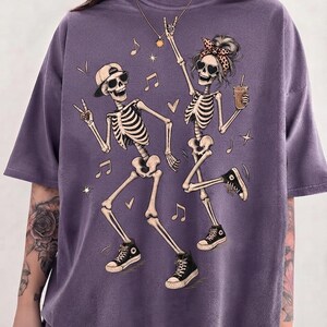 May include: A purple short-sleeved t-shirt featuring a graphic print of two dancing skeletons. One skeleton wears a baseball cap and sunglasses, while the other wears a leopard print headband and sunglasses, holding a drink. Musical notes and hearts surround them.