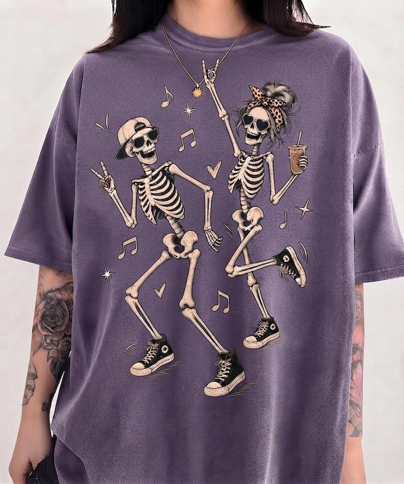 May include: A purple short-sleeved t-shirt featuring a graphic print of two dancing skeletons. One skeleton wears a baseball cap and sunglasses, while the other wears a leopard print headband and sunglasses, holding a drink. Musical notes and hearts surround them.