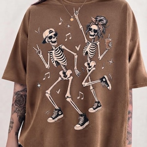 May include: A brown short-sleeved t-shirt featuring a graphic print of two dancing skeletons. One skeleton wears a baseball cap and sunglasses, while the other wears a leopard print headband and heart-shaped sunglasses, holding a beverage cup.