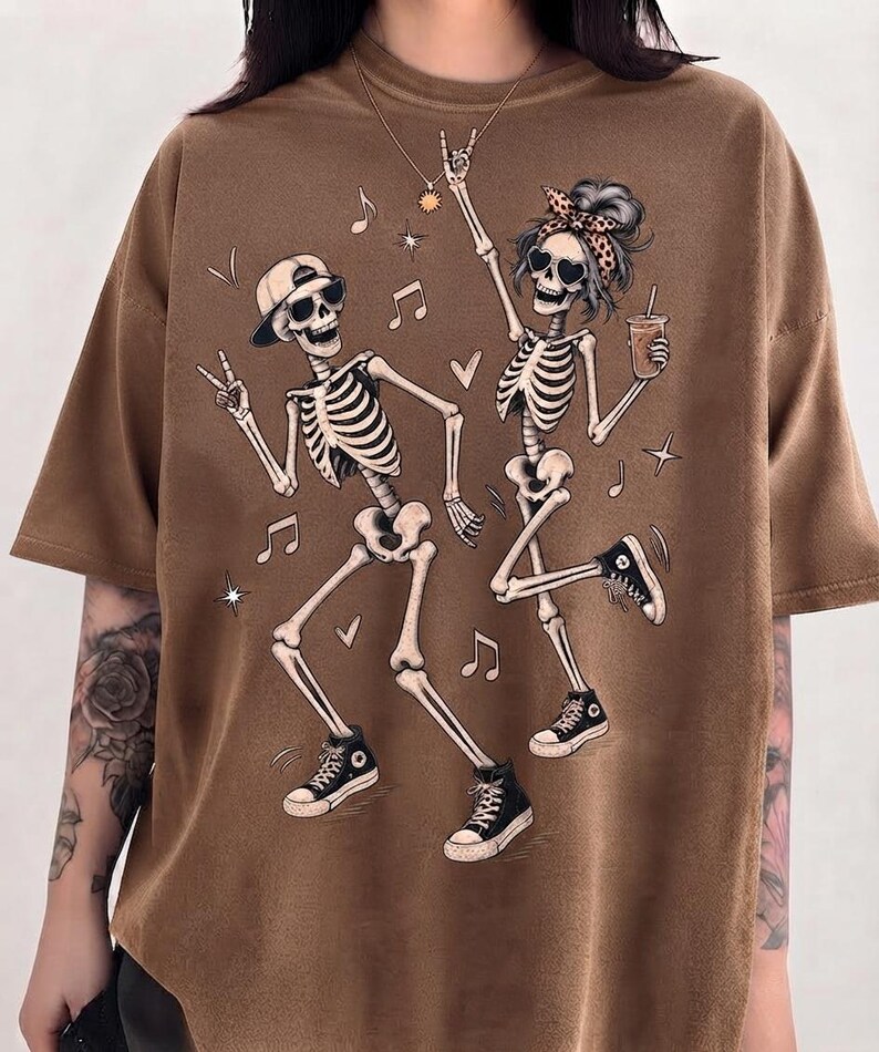 May include: A brown short-sleeved t-shirt featuring a graphic print of two dancing skeletons. One skeleton wears a baseball cap and sunglasses, while the other wears a leopard print headband and heart-shaped sunglasses, holding a beverage cup.