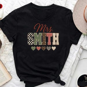 Puede incluir: Camiseta negra con el texto "Mrs. Smith". La palabra "Mrs." está en escritura roja, y "SMITH" está en una mezcla de patrones, incluyendo cuadros, verde, estampado de leopardo y beige. Debajo hay cinco gráficos de corazón.