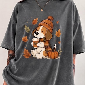Może przedstawiać: Ciemnoszary t-shirt z graficzną ilustracją psa rasy beagle w brązowej czapce z dzianiny i w kratkę szaliku. Pies trzyma kubek kawy z ikoną serca, siedząc obok dyni w otoczeniu opadających jesiennych liści.