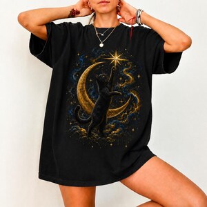 Könnte beinhalten: Schwarzes T-Shirt mit einem Himmelsdesign. Eine schwarze Katze greift nach einem Stern über einem goldenen Halbmond, vor dem Hintergrund blauer und goldener Wolken und Sterne. Das Shirt hat eine bequeme, übergroße Passform.