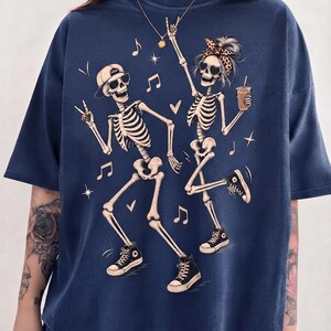 May include: A navy blue t-shirt featuring a graphic print of two dancing skeletons. One skeleton wears a baseball cap and sunglasses, while the other wears a leopard print headband and sunglasses, holding a drink. Musical notes and stars surround them.