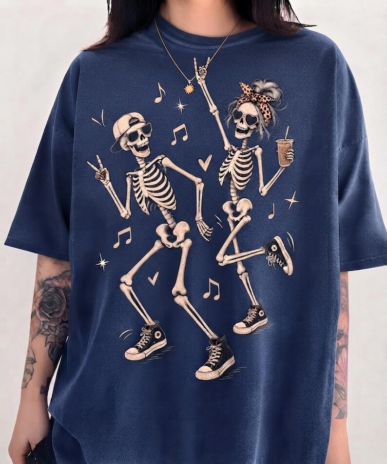 May include: A navy blue t-shirt featuring a graphic print of two dancing skeletons. One skeleton wears a baseball cap and sunglasses, while the other wears a leopard print headband and sunglasses, holding a drink. Musical notes and stars surround them.