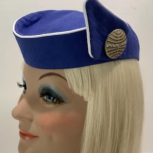 Può includere: Un cappello vintage blu e bianco Pan Am. Il cappello è blu scuro con un bordo bianco e un piccolo bordo arrotondato. Una spilla color oro con il logo Pan Am è fissata sul lato. Il cappello è esposto su una testa di manichino con capelli biondi.
