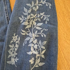 Può includere: Primo piano di jeans blu con un design floreale bianco. Il design presenta foglie e steli dettagliati, creando un motivo botanico. I jeans hanno un orlo grezzo e sono piegati, mostrando l'intricata opera d'arte.