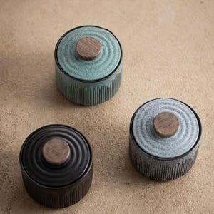 Puede incluir: Tres tarros de cerámica con tapas. Los tarros son en tonos de verde azulado, gris y negro. Cada tarro tiene un pomo de madera en la tapa y detalles acanalados verticales en los lados. Los tarros están sobre una superficie de tono neutro.