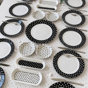 Pode incluir: Um conjunto de louça de cerâmica branca com um padrão de bolinhas pretas nas bordas, incluindo vários pratos redondos, tigelas pequenas e travessas retangulares dispostos sobre uma toalha de mesa a condizer com estampa de bolinhas.