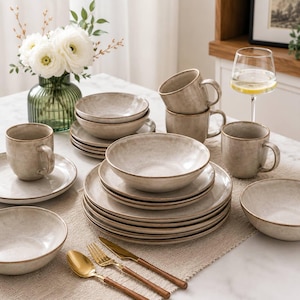 May include: A dinnerware set featuring beige ceramic plates, bowls, and mugs with a subtle speckled finish and brown rims. The collection includes stacked plates and bowls, mugs, and a set of gold-toned flatware with brown handles arranged on a table.
