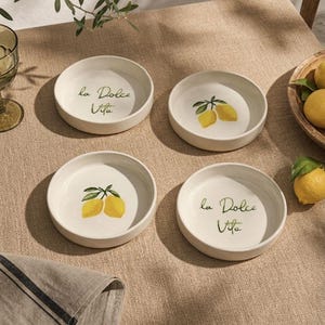 Può includere: Quattro piccole ciotole in ceramica bianca con bordo beige chiaro. Due ciotole presentano un'illustrazione di limone giallo, mentre le altre due hanno la scritta "la Dolce Vita" in verde. Le ciotole sono disposte su una tovaglia beige.