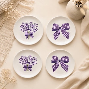 May include: A set of four round white ceramic plates featuring purple illustrations. Two plates display a floral bouquet tied with a gingham bow, while the other two plates feature a single large purple gingham bow design in the center.