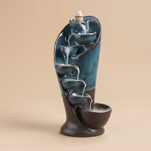 May include: A teal and brown ceramic incense waterfall with a cascading design. The waterfall features multiple tiers of small bowls, with smoke rising from the incense cone at the top. The base is a dark brown color.