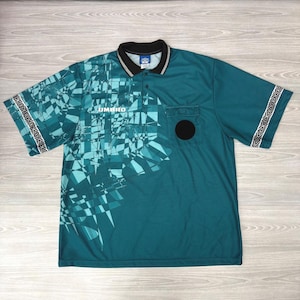 May include: A teal short-sleeve polo shirt featuring a black collar and a geometric pattern on the left side. The shirt includes the white text UMBRO, a black circular patch on the chest pocket, and a diamond-patterned trim on the sleeve cuffs.