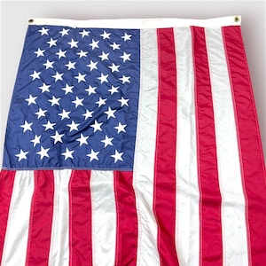 Puede incluir: Una gran bandera estadounidense con un cantón azul lleno de estrellas blancas y rayas rojas y blancas. La bandera está hecha de un material ligero y tiene ojales a lo largo del borde superior para colgar.