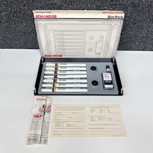 May include: A Koh-I-Noor Rapidograph Slim Pack set. The set includes a variety of white pens with colored tips, ink, and accessories. The packaging is a black and clear plastic case with the brand name prominently displayed.