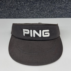 May include: A dark gray visor featuring the word PING embroidered in white capital letters on the front panel. The visor has a curved brim with visible stitching along the edge and a structured crown band that wraps around the head area.