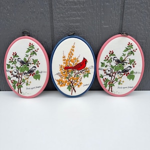 May include: Three oval wall plaques featuring fabric prints. Two pink-framed plaques display Black-capped Chickadees on berry branches. The center blue-framed plaque shows a red cardinal perched on yellow flowering branches. Each has a metal hanger.