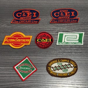 May include: A collection of six embroidered fabric patches featuring various railroad logos. The patches include Speed C&EI Service, Alton & Southern, C&EI Chicago and Eastern Illinois, Penn Central, American Railway Express, and Springs & Cripple Creek.