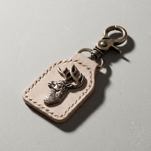 May include: A light beige leather keychain featuring a rectangular tag with visible stitching along the edges. A bronze-toned metal emblem of a deer head with antlers is centered on the leather. The tag is attached to a bronze-toned metal lobster clasp.