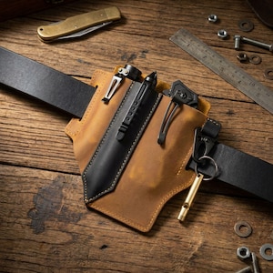 May include: A tan leather belt sheath featuring a black leather overlay, attached to a black leather belt. The sheath holds a black metal pen and a black metal tool with a clip. A brass whistle hangs from a black metal ring on the side.