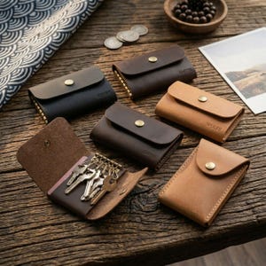 Könnte beinhalten: Eine Sammlung von Schlüsseletuis aus Leder in Schwarz, Dunkelbraun und Hellbraun, jeweils mit einem goldfarbenen Druckknopfverschluss. Ein Etui ist geöffnet und zeigt einen Metallschlüsselhalter mit mehreren Schlüsseln. Einige Etuis tragen Namen.
