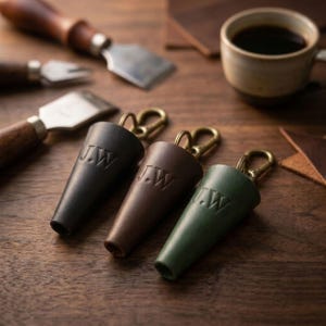 May include: Three cone-shaped leather keychains in black, dark brown, and forest green, each featuring the embossed initials J.W. and a brass-toned metal lobster clasp. The keychains are arranged in a row on a wooden surface near leather tools.